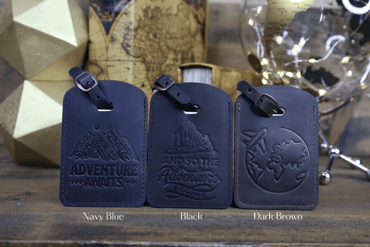 Three leather luggage tags in navy blue, black, and dark brown with embossed designs on a wooden surface.